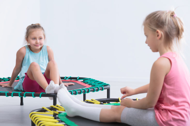 Two little girls sitting on trampolines in a very bright room, smiling at each other